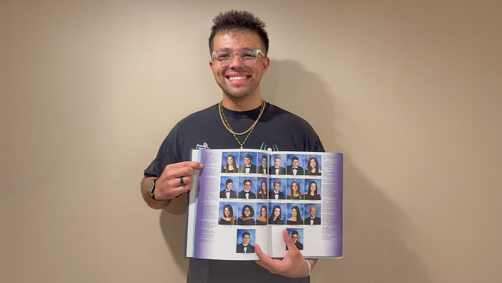 A young man wearing glasses, holding an open yearbook and smiling