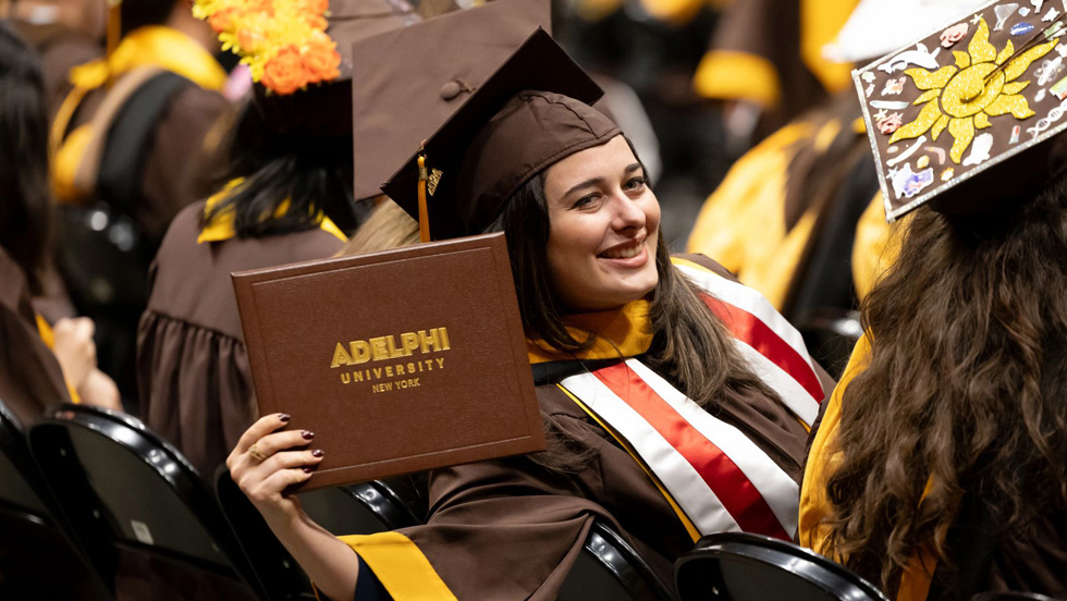 Halpert, in cap and gown, displays her diploma while seated in a row with fellow graduates.