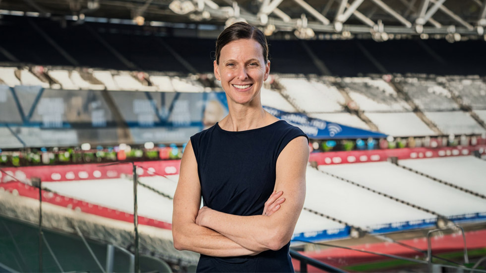 A professional headshot of a woman wearing business attire and smiling with her arms crossed in front of a sports arena