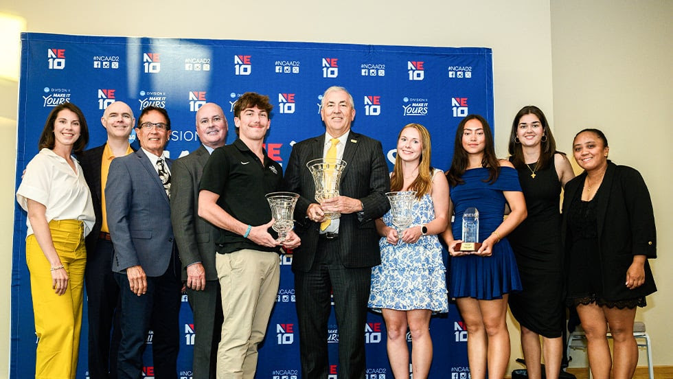 Surrounded by award-winning athletes, coaches and University leadership, Adelphi's new president, Michael A.L. Balboni '81, JD, center, proudly holds the 2026 Presidents Cup.