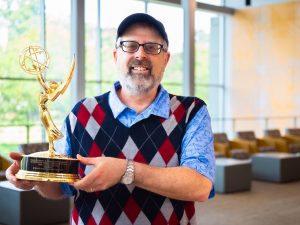Professor Shore, wearing his customary argyle vest and standing inside Adelphi's University Center building, holds the Emmy trophy.