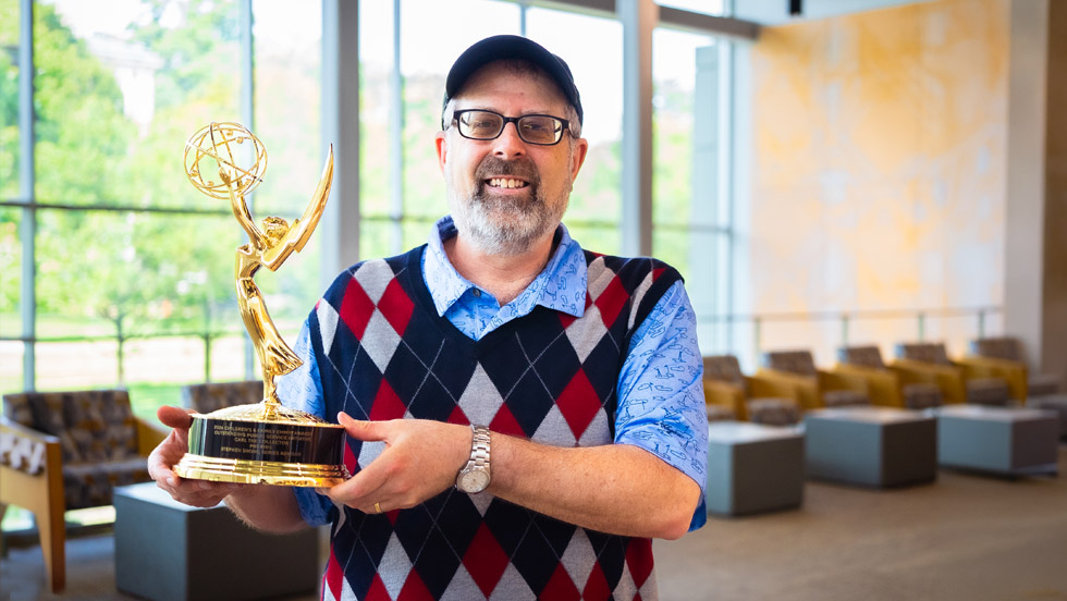 Professor Shore, wearing his customary argyle vest and standing inside Adelphi's University Center building, holds the Emmy trophy.