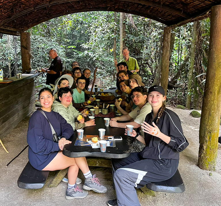 A large group of students and their professors eat lunch on a picnic beach, under a wooden shelter.