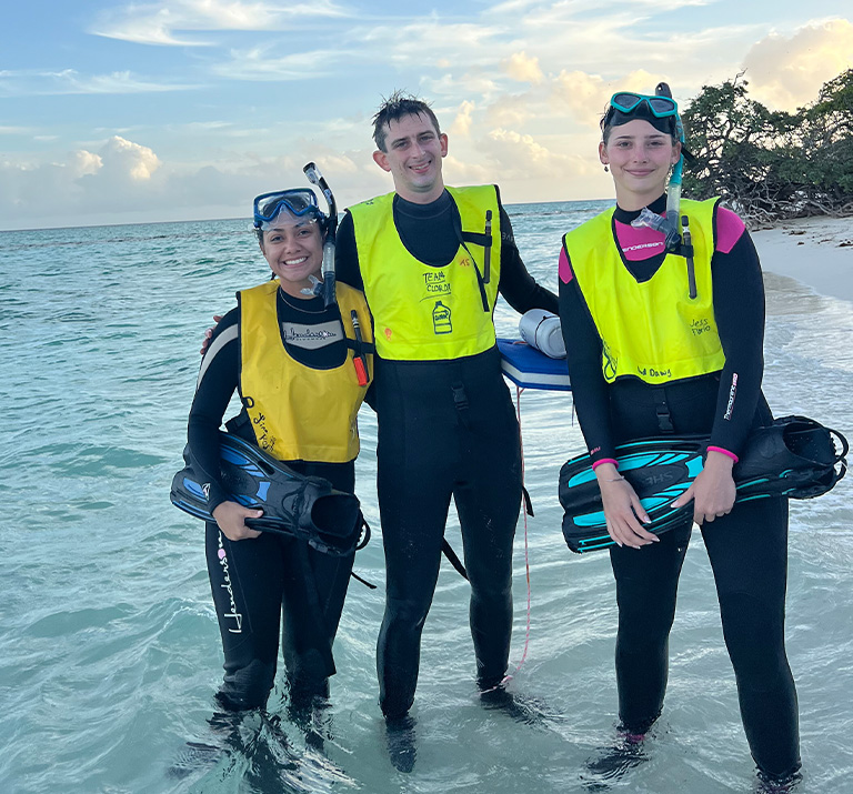 A male student and two female students in full scuba gear pose along a sandy white beach.