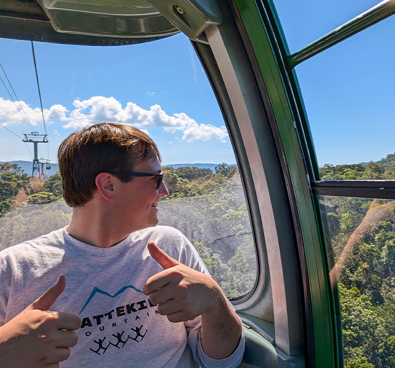 A male student looks down from a gondola high above a green forest.