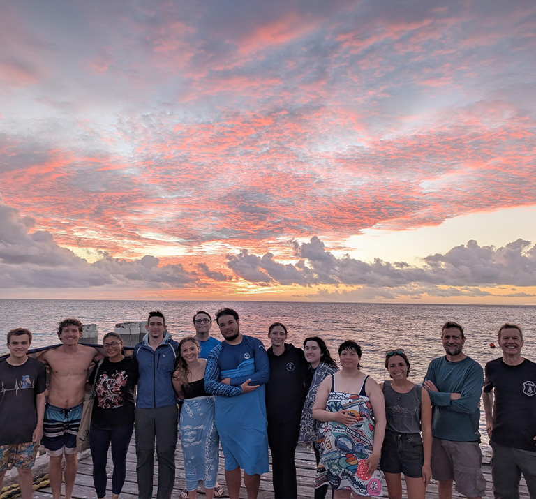 A row of 13 students and professors stand along a sandy coast in front of a pink sunset.