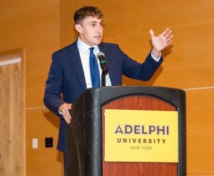A young man in a suit and tie gestures at a an Adelphi podium.