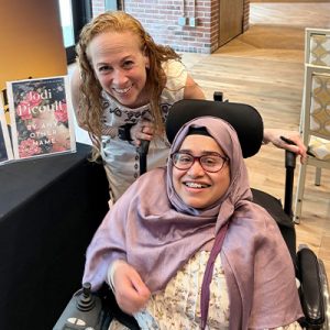 Jodi Picoult bends down to pose with Masud. She is standing next to a copy of her novel By Any Other Name.