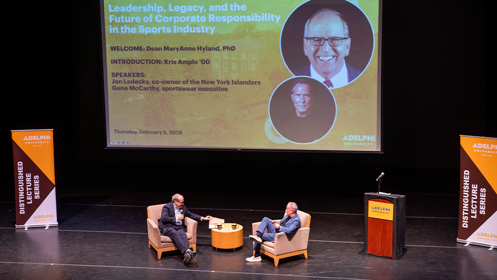 Two men sit in armchairs on a stage, talking, with a low table and coffee cups between them. Behind them, a large screen displays the event title "Leadership, Legacy, and the Future of Corporate Responsibility in the Sports Industry," hosted by Adelphi University's Distinguished Lecture Series, dated Thursday, February 5, 2026, alongside photos of the two speakers. A podium with a microphone stands to the right, and branded pull-up banners reading "Distinguished Lecture Series" flank the stage on both sides.