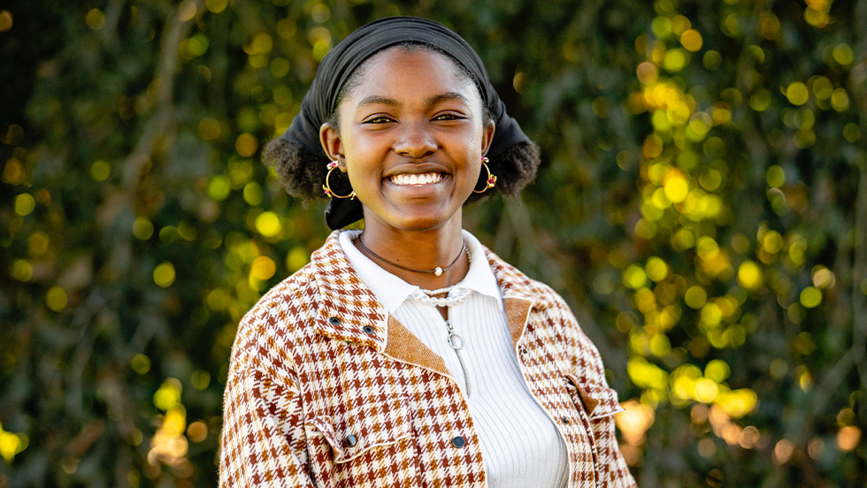 A young Black female student smiles broadly, surrounded by greenery on Adelphi's campus