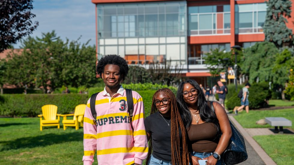 A group of three students smiling in front of a modern orange university building with a walkway and trees in the background