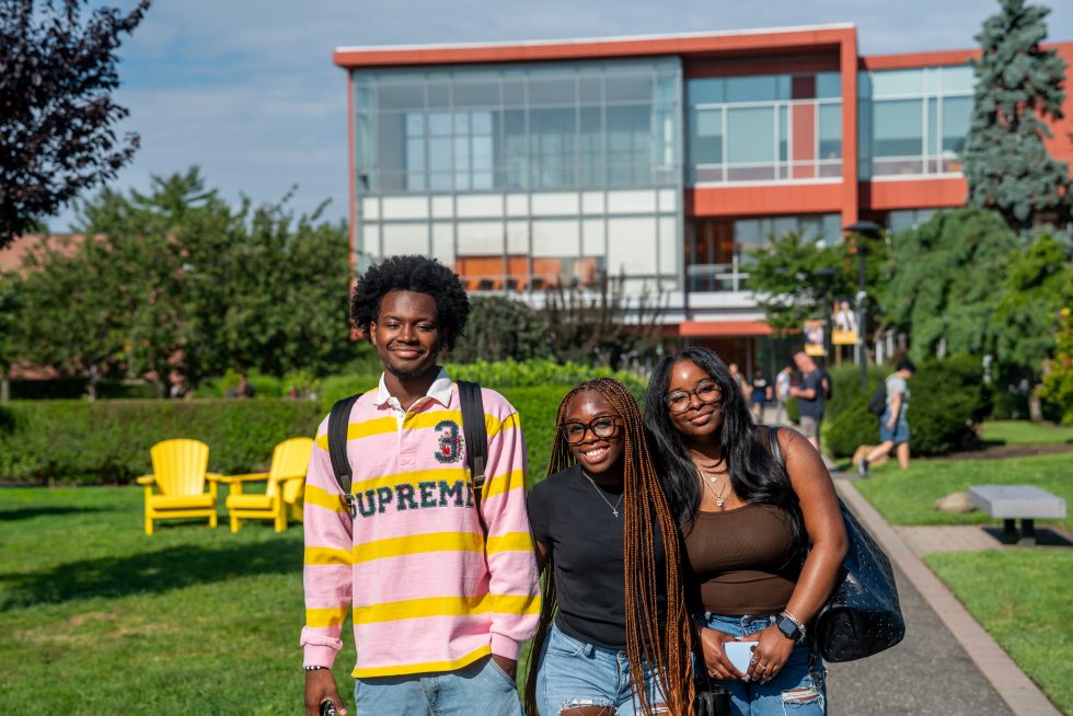 A group of three students smiling in front of a modern orange university building with a walkway and trees in the background
