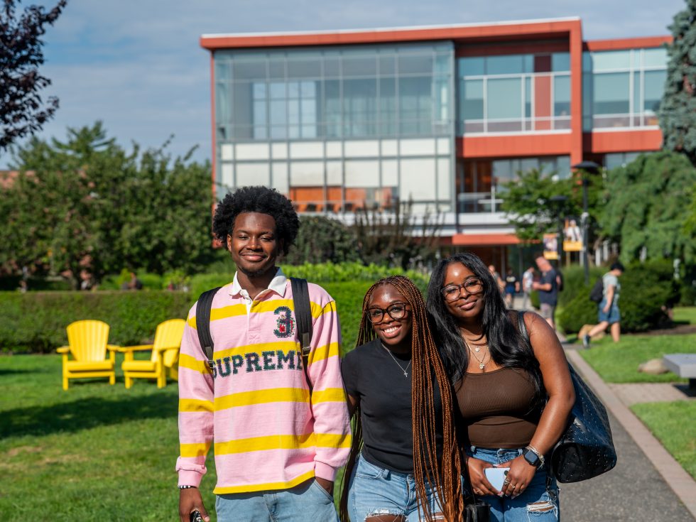 A group of three students smiling in front of a modern orange university building with a walkway and trees in the background