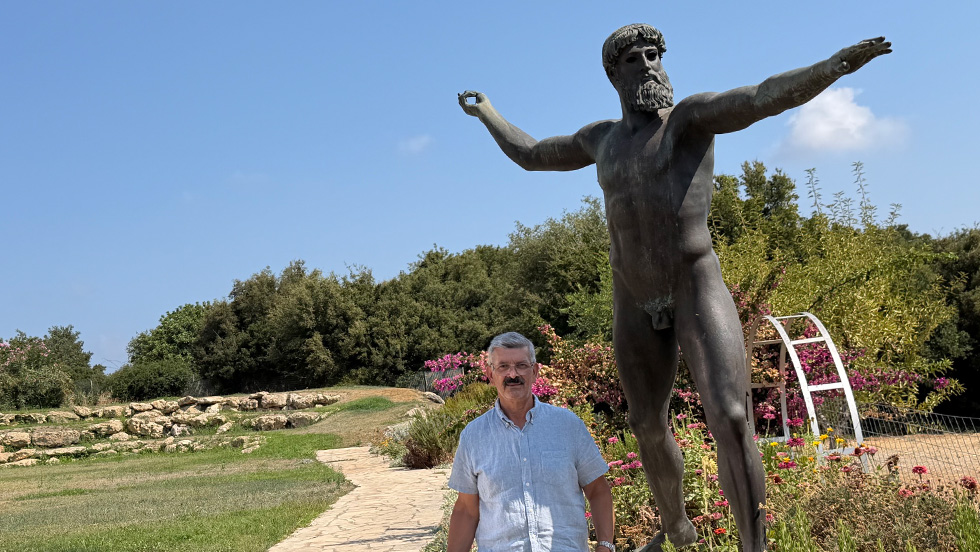 Professor Agelarakis in a short-sleeve shirt on a brilliantly sunny day, next to the statue. The statue depicts a naked figure, considered to be either Zeus or Poseidon, pointing toward nearby Mount Ida. 