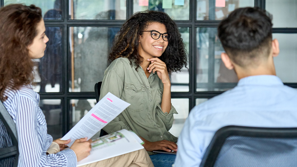 A woman with curly hair and glasses smiles while seated in an office meeting, resting her chin on her hand. Across from her, two colleagues sit with their backs partially to the camera — one holding a document with a pink highlighted section, the other in a light blue shirt. A glass-walled office with sticky notes is visible in the background.