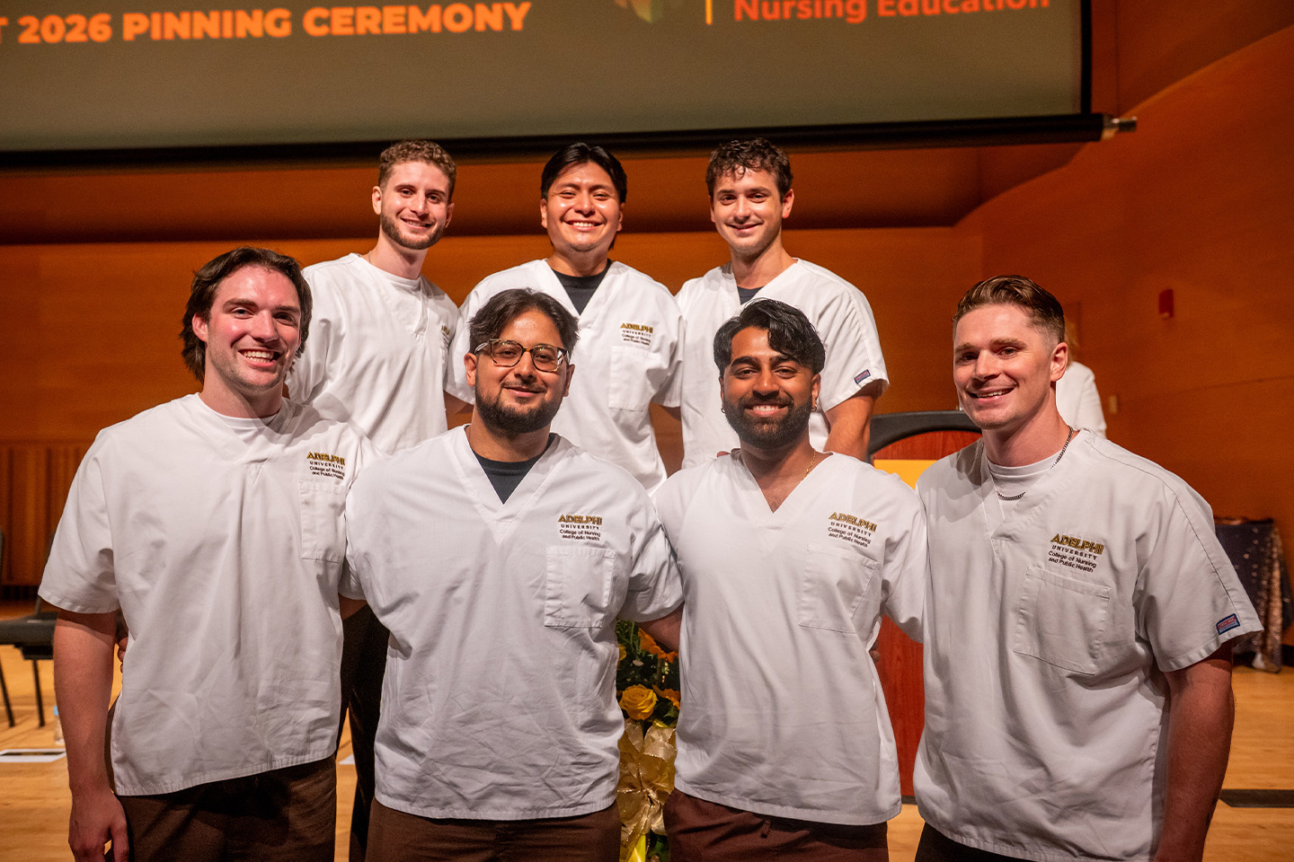 Seven male nursing students beam proudly, wearing their nursing uniforms.