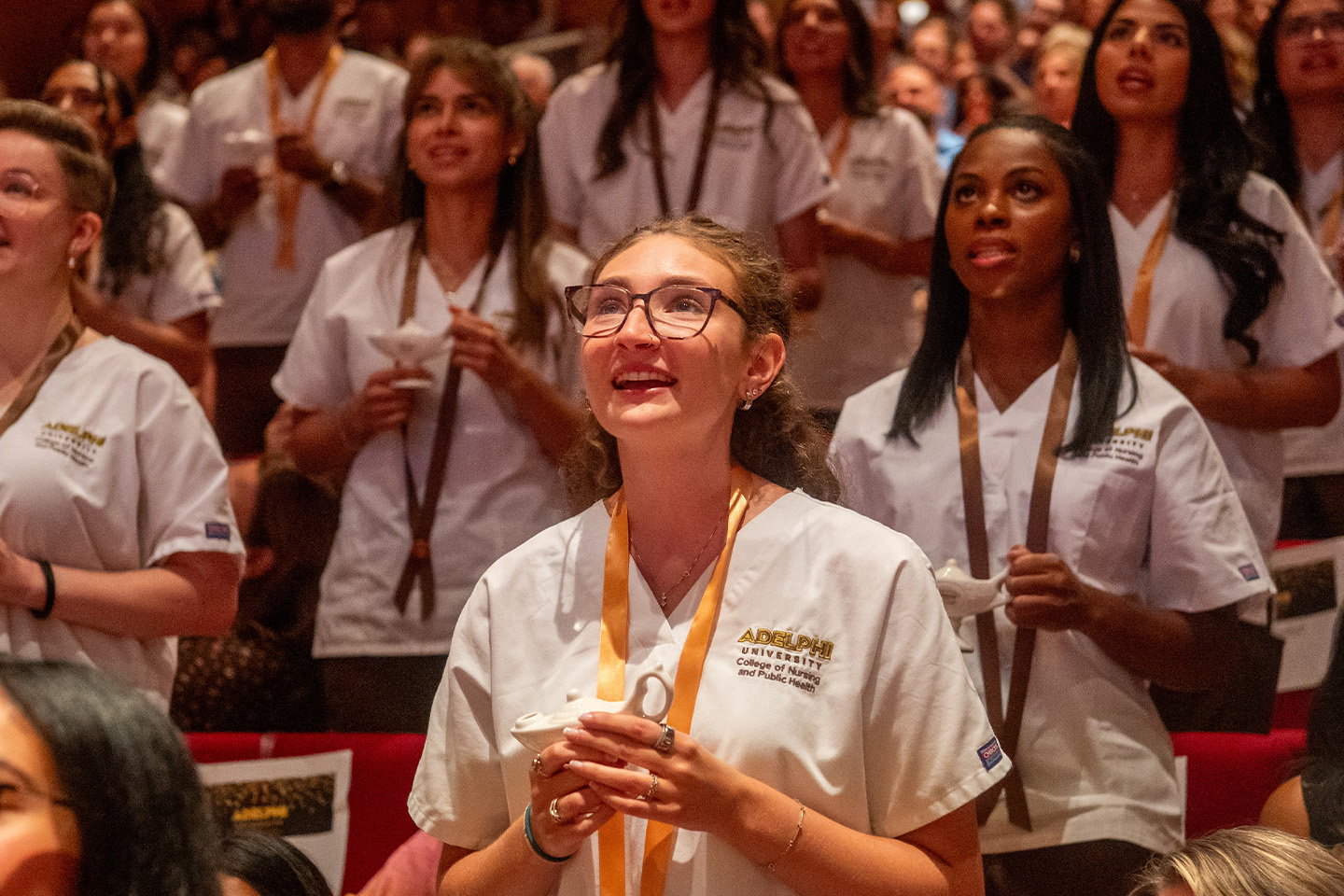 A large group of students in nursing uniforms hold mini lamps and recite a pledge.