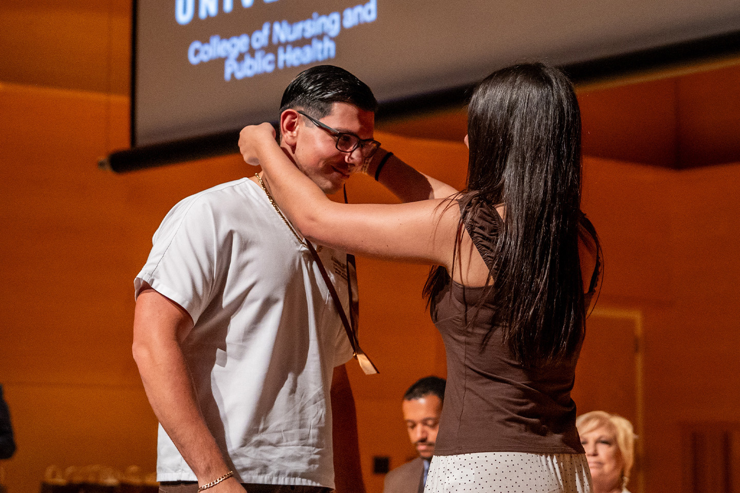 A male nursing grad, with a happy smile, receives a ribbon and pin from a woman as staff look on.