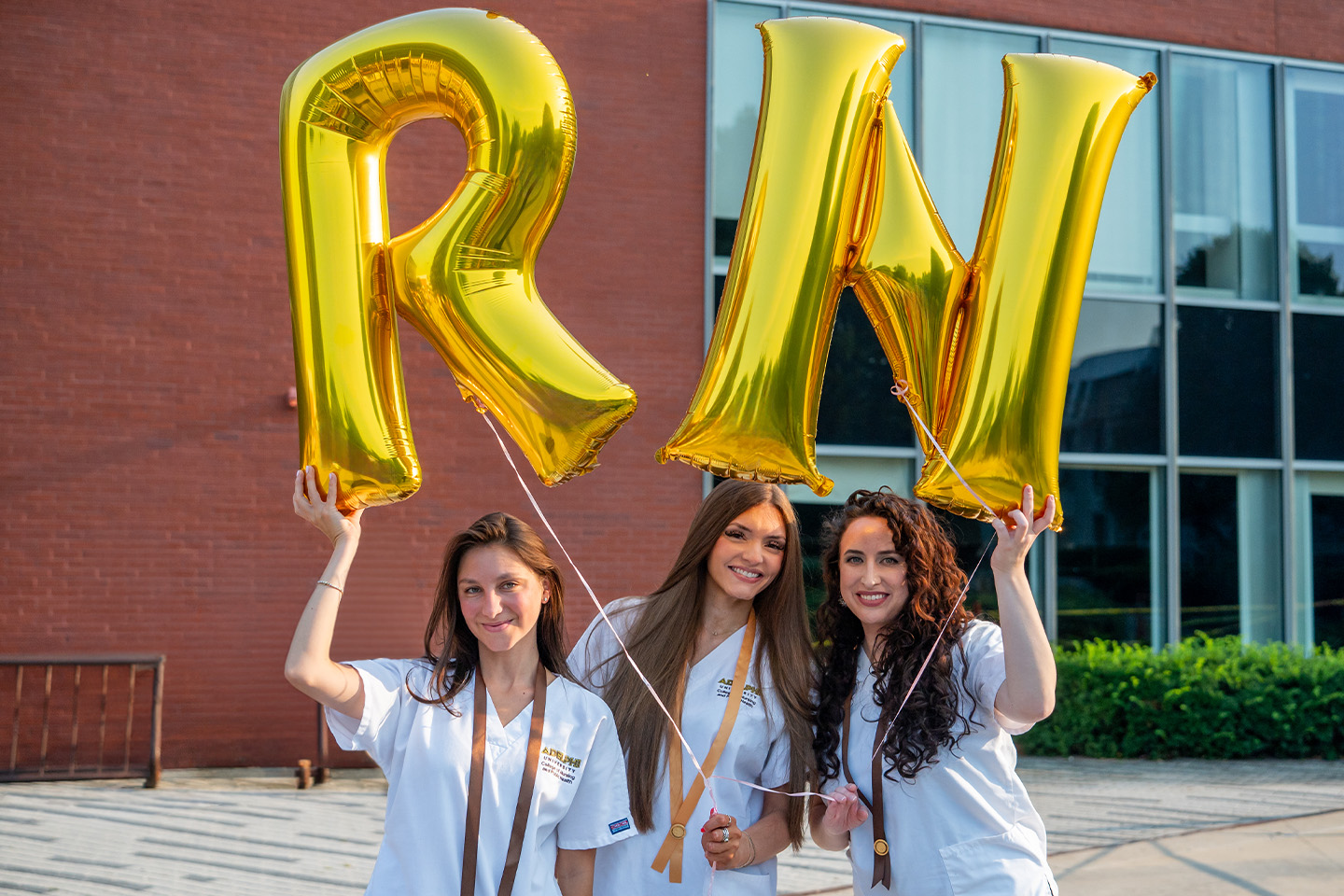 Three women grads hold Mylar balloons that spell RN.