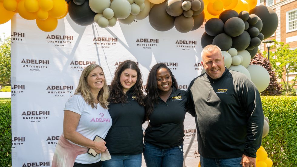 A group of four people smiling in front of an Adelphi-branded step and repeat background with balloons.