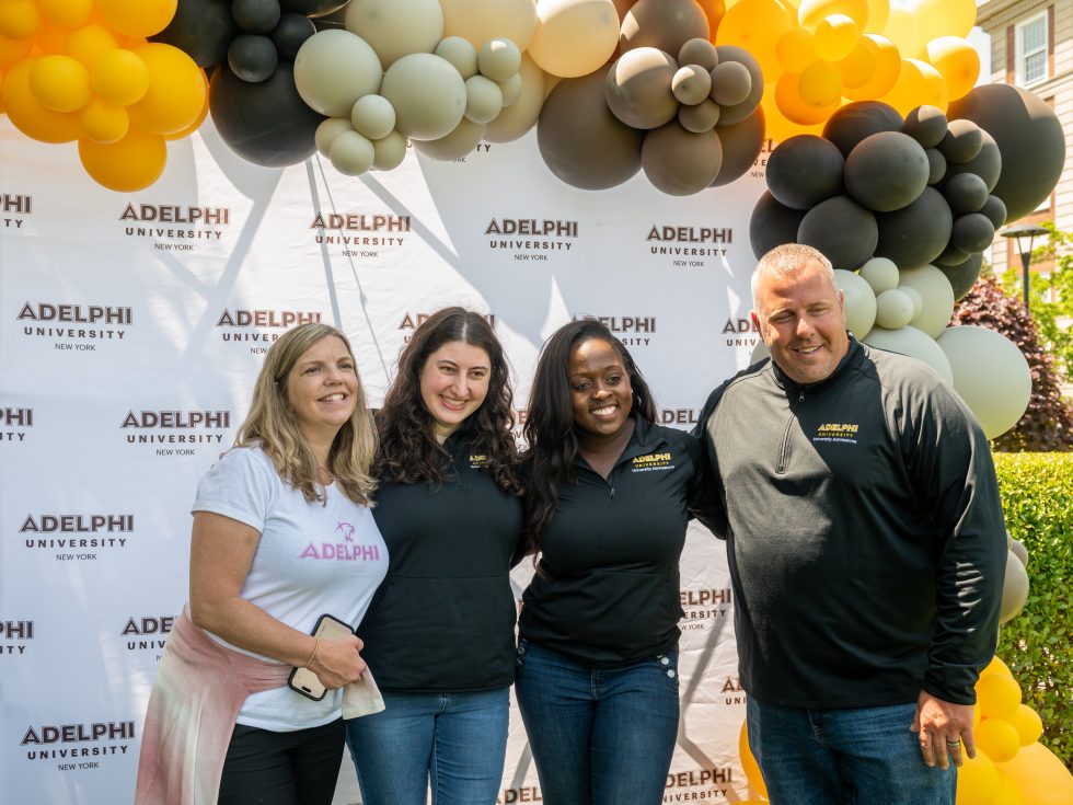 A group of four people smiling in front of an Adelphi-branded step and repeat background with balloons.