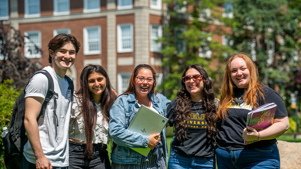 A group of five Adelphi students smiling in front of a university building holding laptops and books