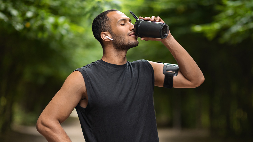 A man in workout gear pauses on a tree-lined path to drink from a shaker bottle, wearing an armband phone holder and wireless earbuds.