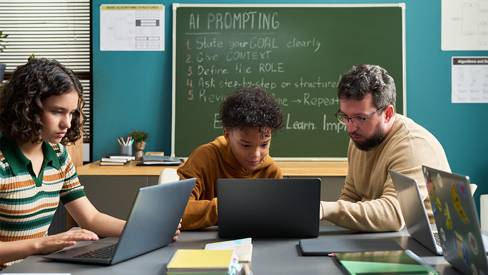 A teacher helps two students on laptops in a classroom with a chalkboard listing steps for AI prompting: state your goal, give context, define the role, ask step-by-step, and revise.