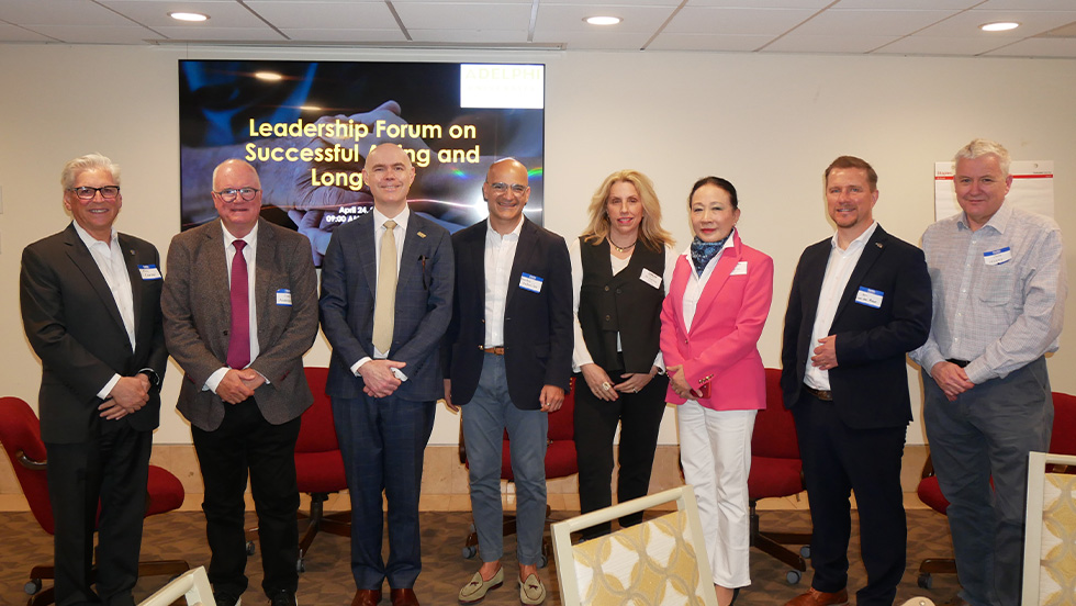 A group of eight professionals in business attire pose together in a conference room in front of a screen displaying "Leadership Forum on Successful Aging and Longevity."
