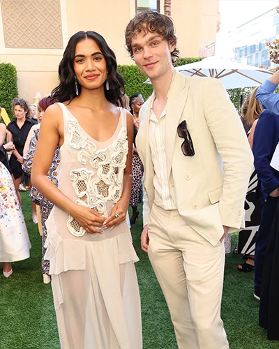 At a festive outdoor party, a young man in a white suit stands by a dark-haired actress in a white gown.