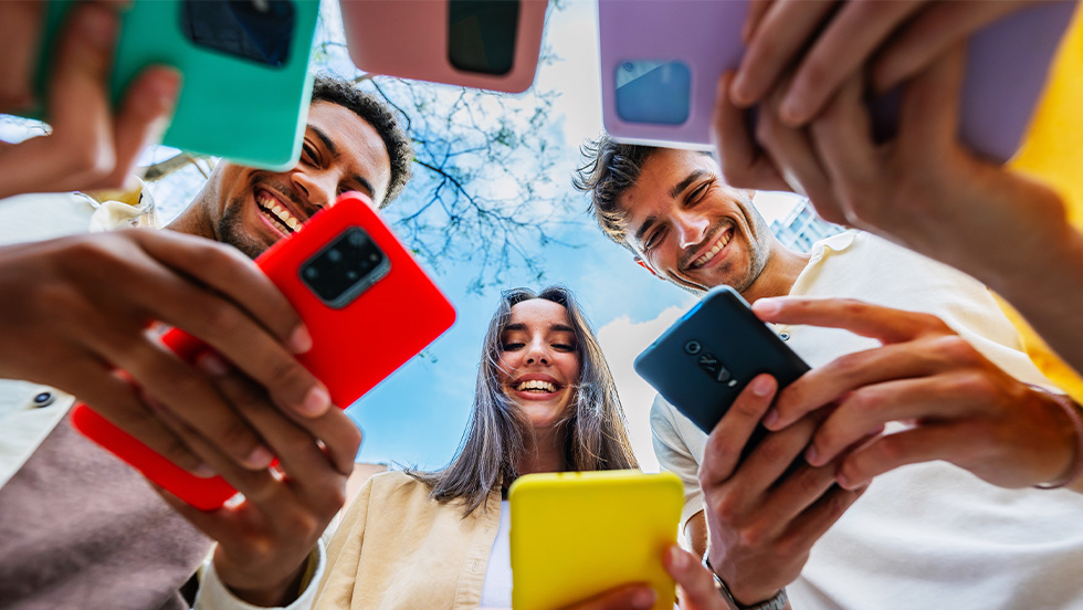 A group of friends stand in a circle holding colorful smartphones above their heads, photographed from below looking up. They smile down at the camera with blue sky and bare tree branches visible in the background.