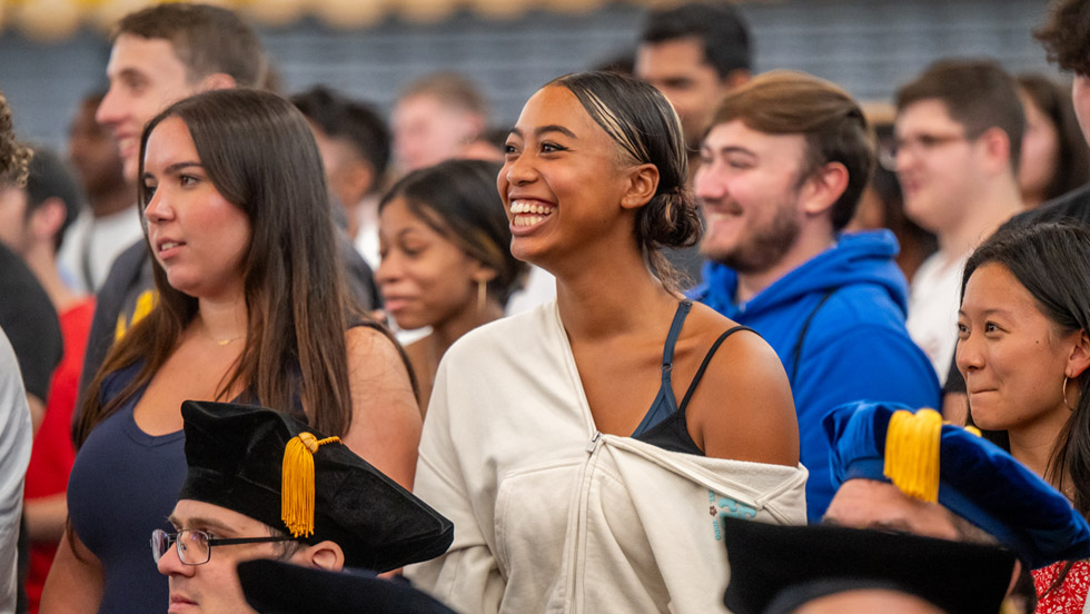 Smiling students amid the crowd of fellow first-year classmates, with faculty members in cap and gown seated in front of them