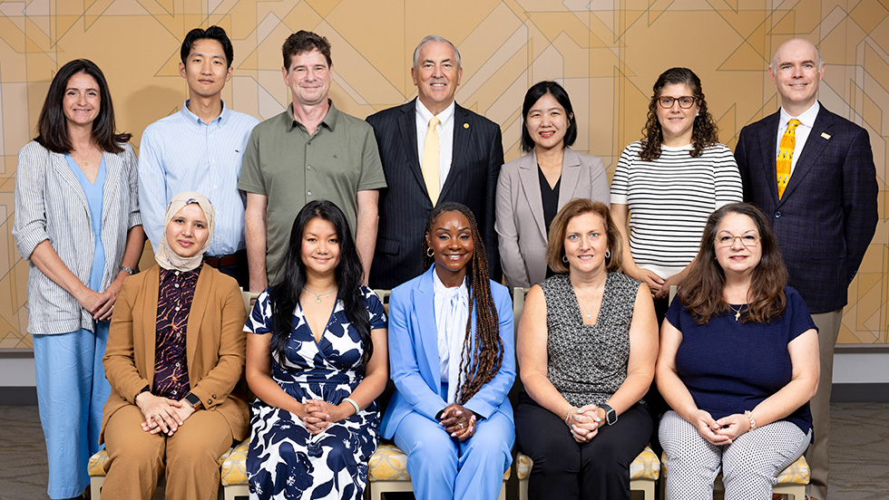 Group portrait of twelve new faculty members and a university administrator, arranged in two rows against a gold patterned wall.
