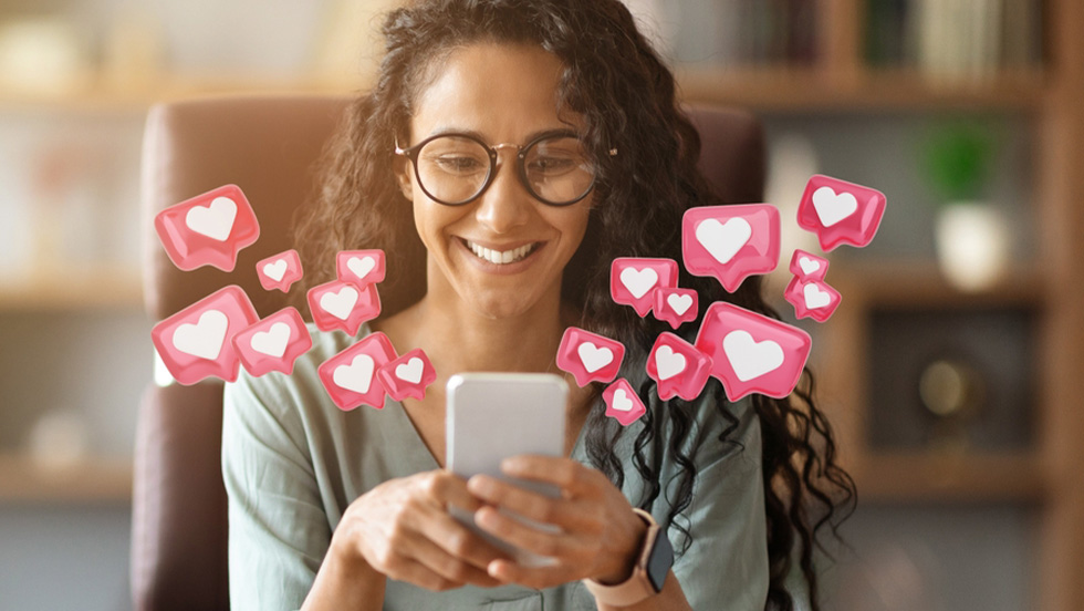 A smiling woman with glasses and long, dark curly hair looks at her smartphone while 3D pink heart notification bubbles float around the screen against a warm background.
