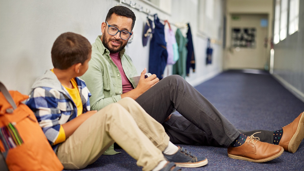 A male school psychologist wearing glasses sits on the carpeted floor of a school hallway, speaking attentively with a young boy sitting beside his orange backpack.