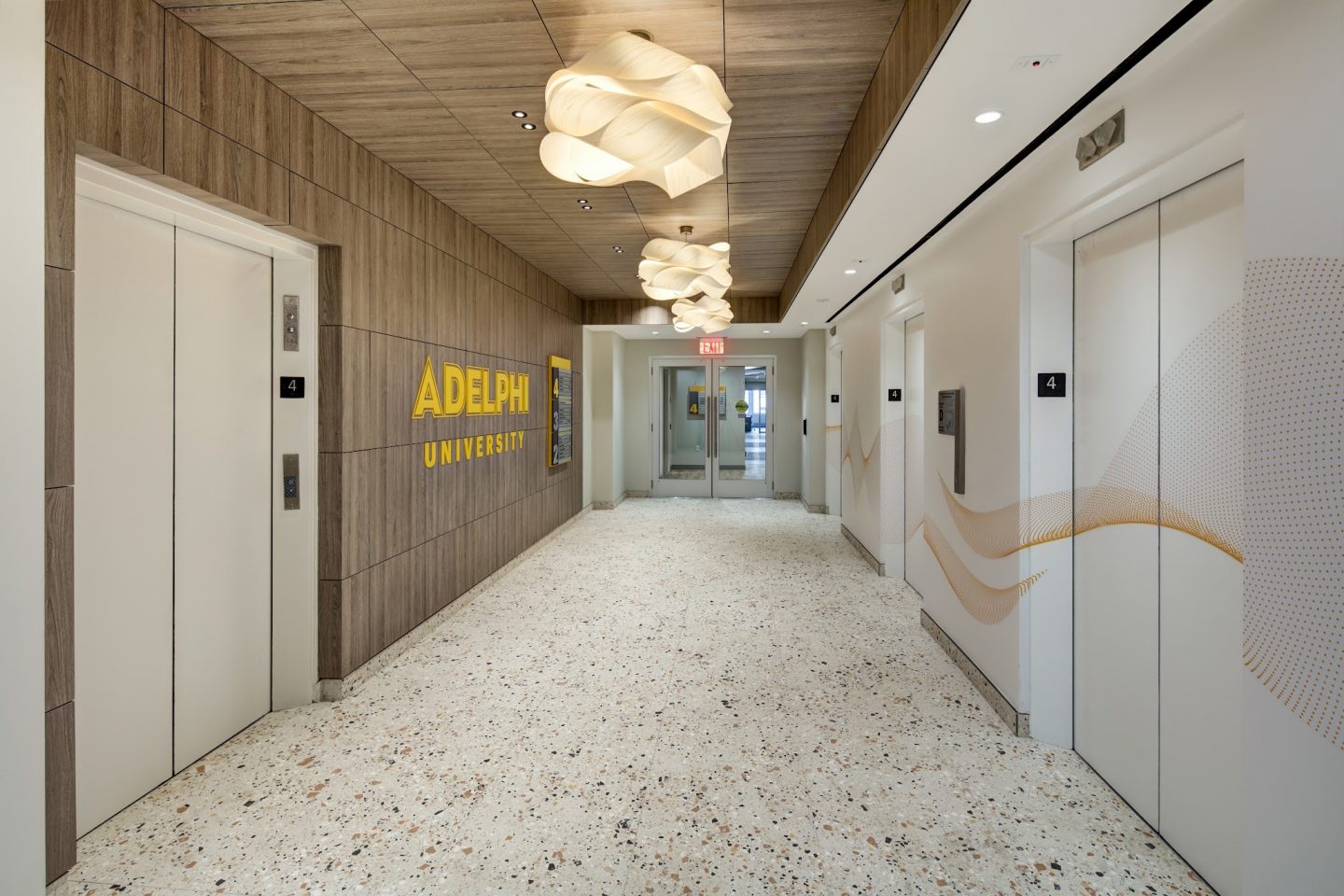 An empty elevator lobby with wood-paneled walls, a yellow “ADELPHI UNIVERSITY” wall sign, layered wave-shaped pendant lights hanging from a wood-slat ceiling, white elevator doors on both sides, and a speckled terrazzo floor leading to glass entry doors marked with a floor number “4”