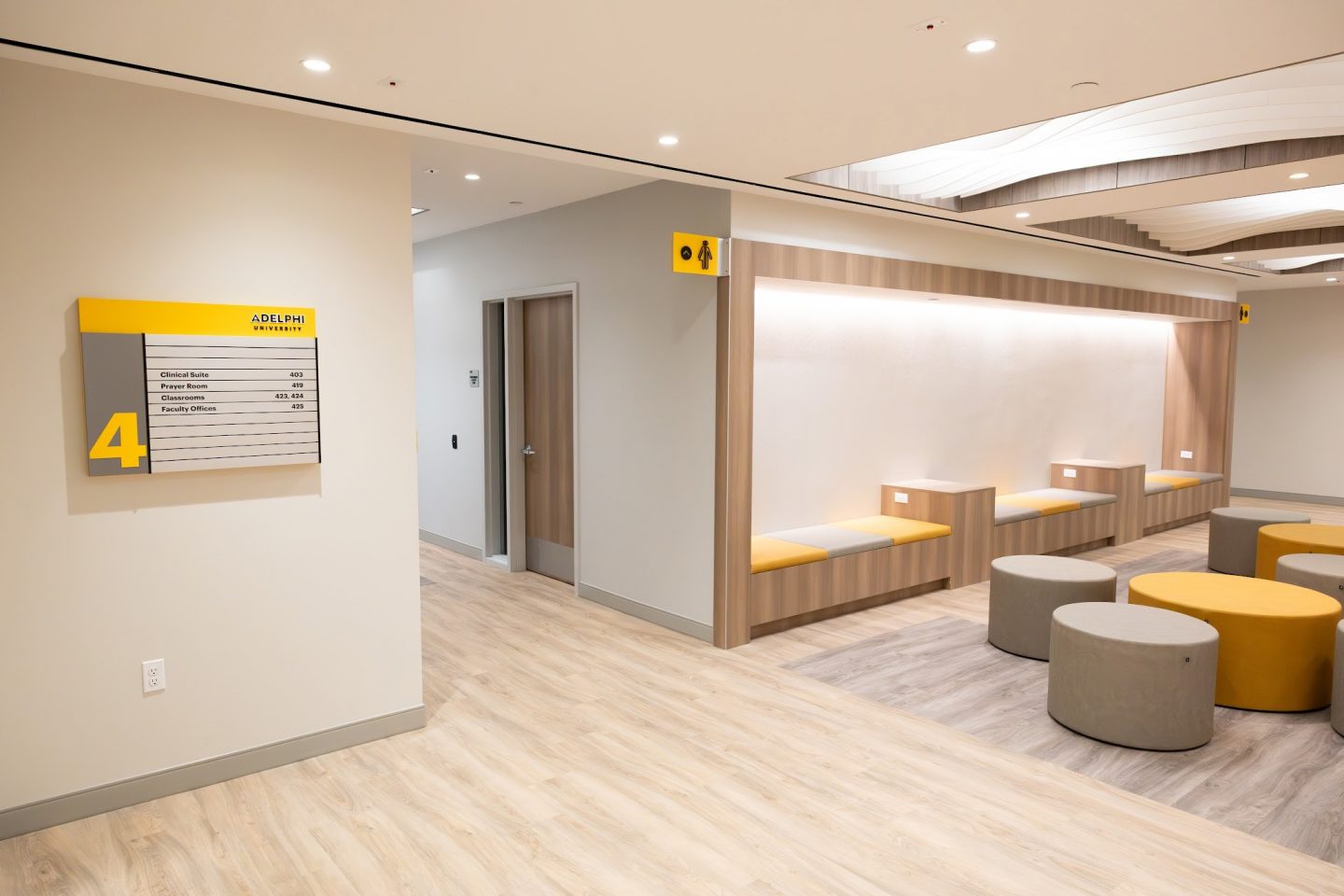 Hallway with a yellow-and-gray “4” floor directory sign listing Clinical Suite 403, Prayer Room 419, Classrooms 423/424 and Faculty Offices 425, next to a wood-paneled alcove with built-in bench seating and round ottomans on light wood flooring