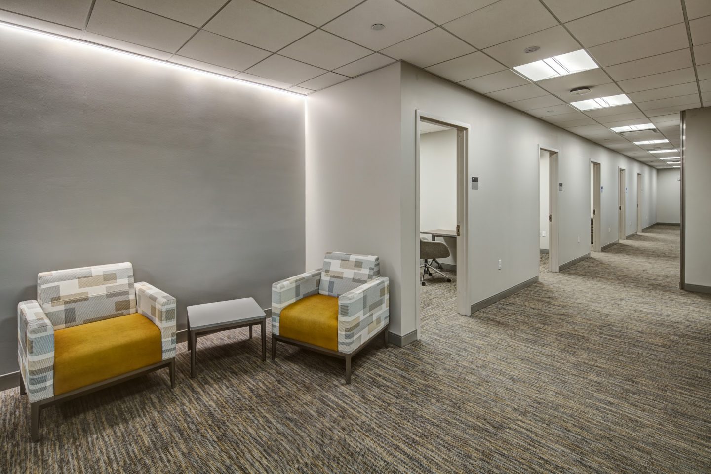 A hallway waiting area for the Derner Center for Psychological Services clinical suites, with two patterned chairs featuring mustard-yellow seat cushions, a small side table and a row of private office doors extending down a carpeted corridor