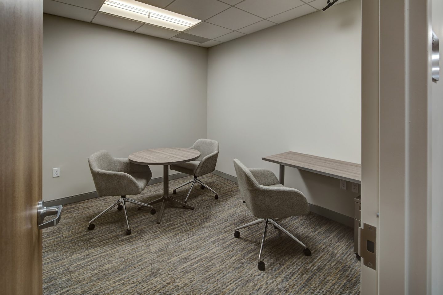 A small treatment room with pale walls and patterned carpet, furnished with a round table and two upholstered swivel chairs, a third matching chair near a built-in desk along the side wall, viewed through an open door