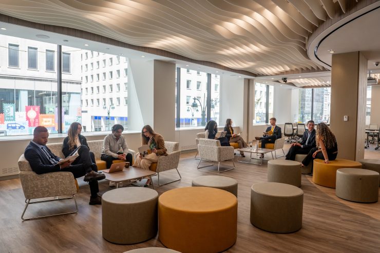 A bright open lounge with wood-slat wave ceiling, floor-to-ceiling windows showing city buildings, and several students in business casual attire sitting on grey and mustard-yellow chairs and round ottomans, reading and chatting