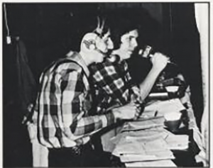A DJ talks into a microphone, with an engineer by his side at a cluttered desk.