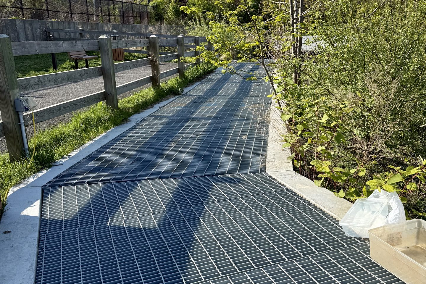 A metal grate walkway/bridge over a drainage channel, flanked by a wooden split-rail fence on the left and dense green shrubs and a young tree on the right, with utility poles and power lines visible in the sky above. A clear storage bin sits on the grating in the foreground.