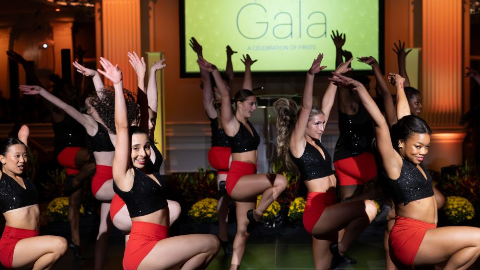 Dancers in black tops and red shorts perform with arms raised in a ballroom, with an Adelphi University Gala screen behind them.