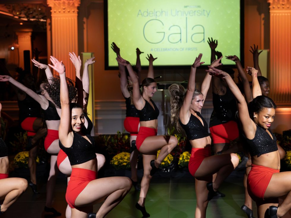 Dancers in black tops and red shorts perform with arms raised in a ballroom, with an Adelphi University Gala screen behind them.