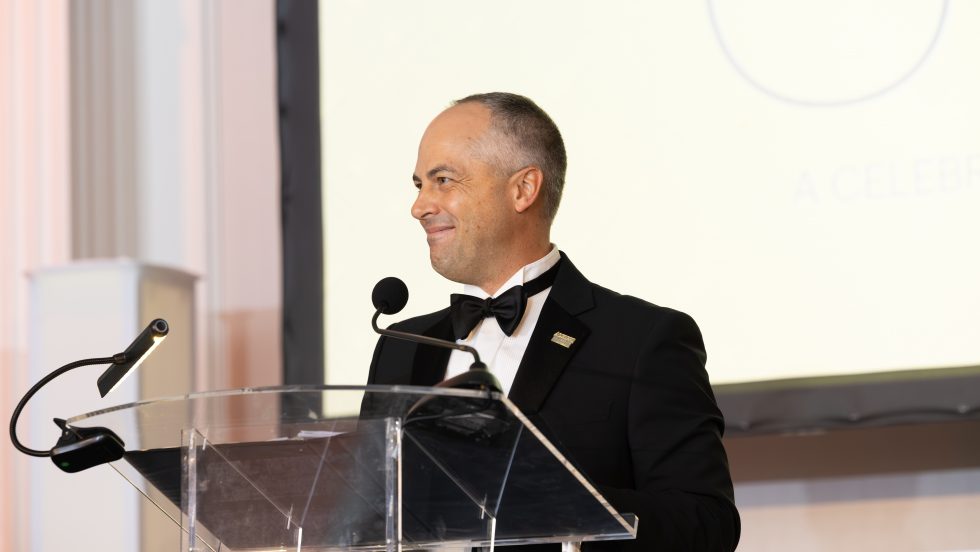 Thomas A. Kline, wearing a black tuxedo, applauds behind a clear lectern at Adelphi’s 2026 Gala.