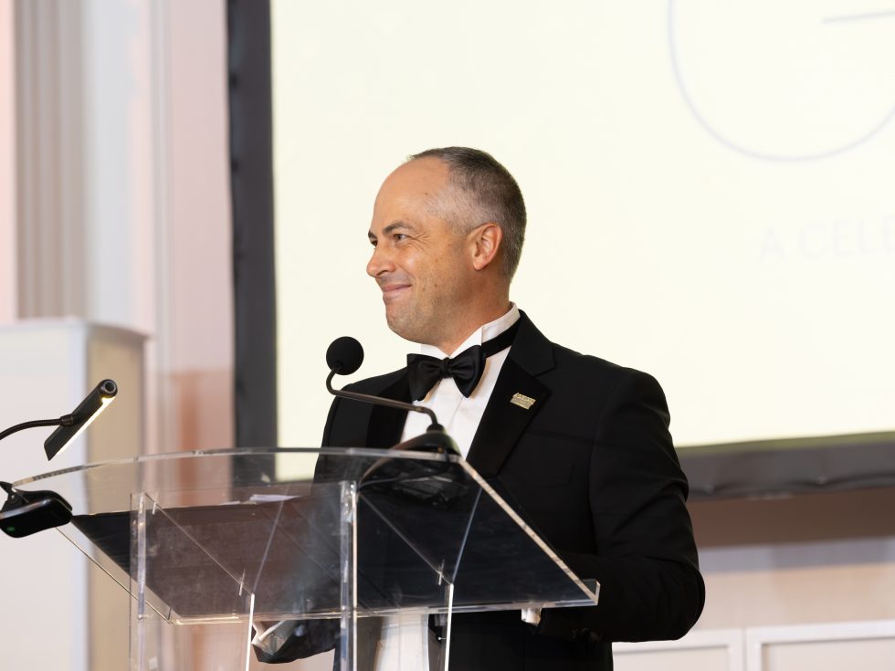Thomas A. Kline, wearing a black tuxedo, applauds behind a clear lectern at Adelphi’s 2026 Gala.