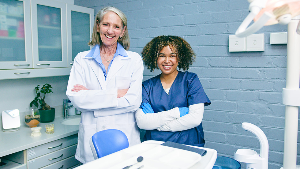 Two smiling healthcare professionals stand side by side in a dental treatment room with their arms crossed. An older woman with shoulder-length gray hair wears a white lab coat, and a younger woman with curly hair wears navy blue scrubs and blue gloves. A dental chair, overhead light, and cabinets are visible behind them.