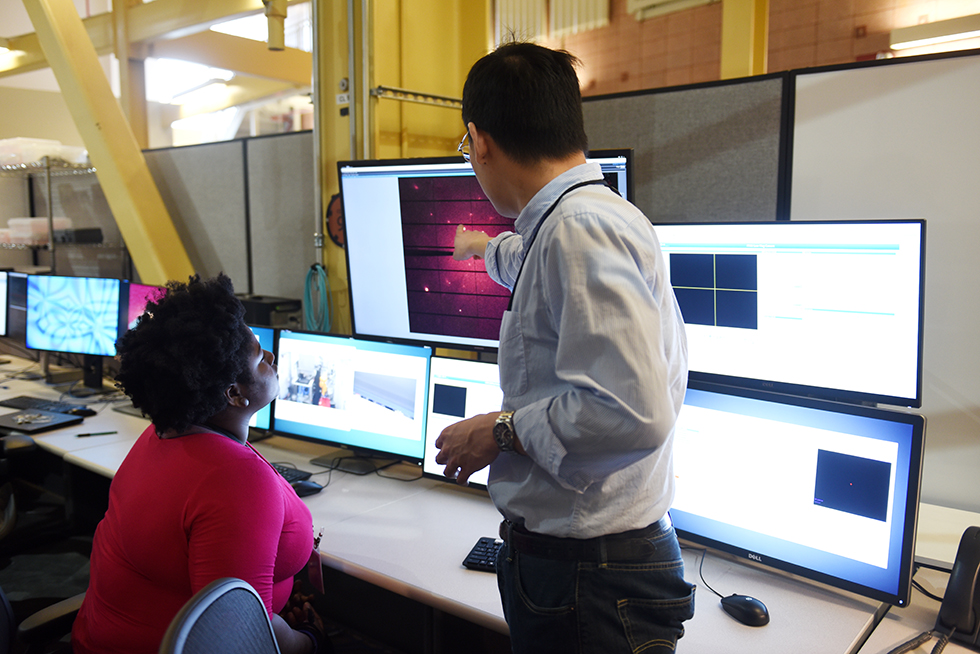 A student sits at a table looking at a computer screen as a scientist points out something on the screen.