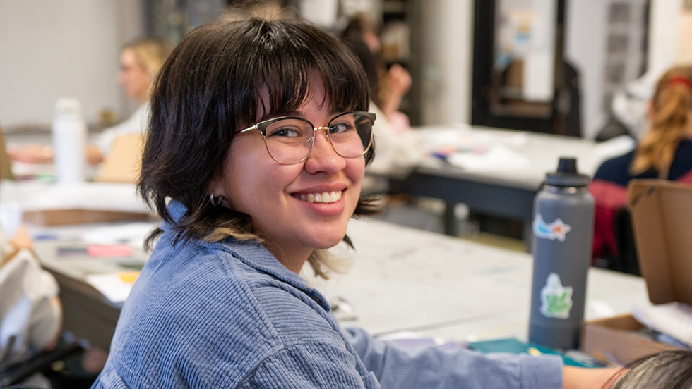 A young student smiles as she turns from a classroom desk to fact the camera.