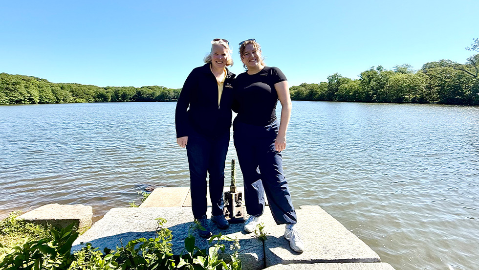 Two women stand together on a stone platform at the edge of a lake on a sunny day, smiling for the camera. Both wear sunglasses pushed up on their heads and dark clothing. Calm water and a tree-lined shoreline stretch behind them under a clear blue sky.